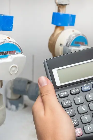 Mão fazendo contas na calculadora em frente a dois hidrômetros